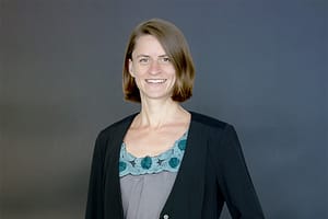 Dr. Ruth Maria Schüler steht vor dunklem Hintergrund und lächelt freundlich in die Kamera.