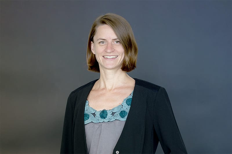 Dr. Ruth Maria Schüler steht vor dunklem Hintergrund und lächelt freundlich in die Kamera.